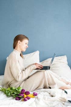 Happy smiling woman sitting on the bed wearing pajamas, with pleasure enjoying flowers, chatting using laptop