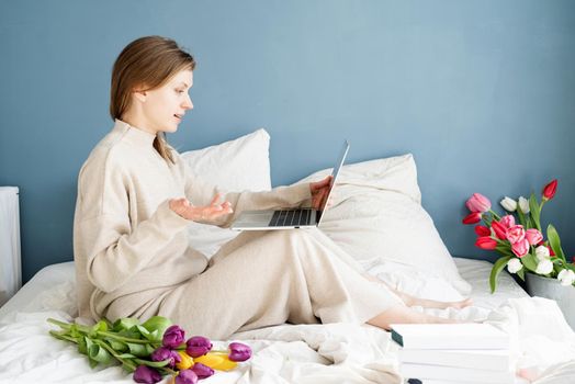 Happy smiling woman sitting on the bed wearing pajamas, with pleasure enjoying flowers, chatting using laptop