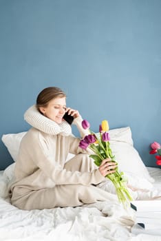 Happy woman sitting on the bed wearing pajamas talking on the phone, blue wall background