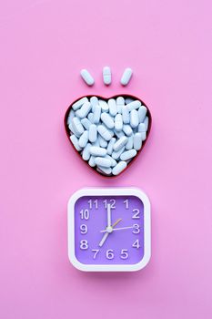Clock with medical blue pills in heart shaped box on pink background. Top view