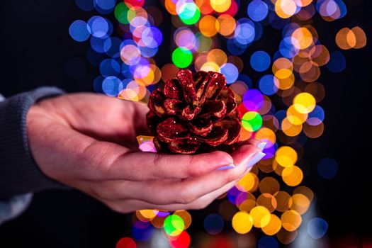 Holding Christmas pine cone decoration isolated on background with blurred lights. December season, Christmas composition.