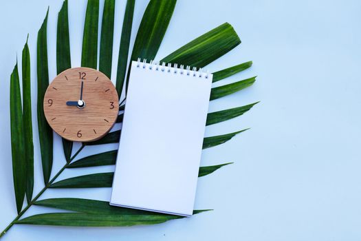 Wooden clock with notebook on green leaves on white background. Copy space