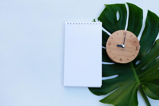 Wooden clock with notebook on green leaves on white background. Copy space