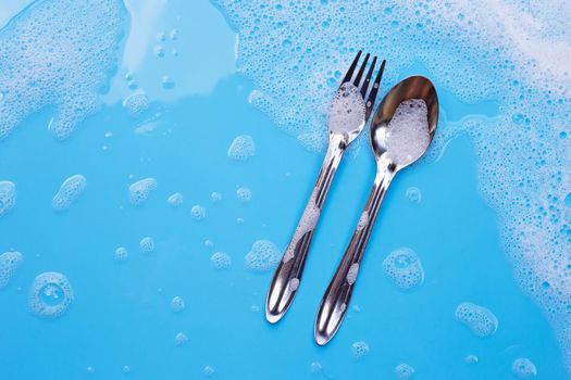 Washing dishes concept. Fork with spoon on wet blue background with soapy foam 