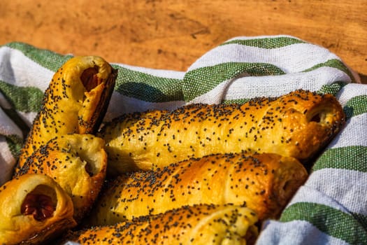 Sausages baked in dough sprinkled with salt and poppy seeds in a rustic basket. Sausages rolls, delicious homemade pastries in a rustic composition.