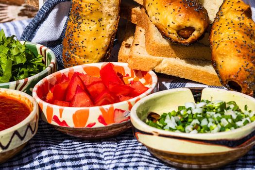 Rustic composition with sausages rolls and different bowls with sauce and chopped vegetables ( tomatoes, green lettuce, green onion, green garlic)