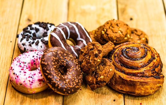 Colorful donuts, puff pastry and biscuits on wooden table. Sweet icing sugar food with glazed sprinkles, doughnut with chocolate frosting. Top view with copy space