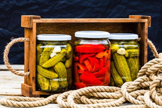 Wooden crate with glass jars with pickled red bell peppers and pickled cucumbers (pickles) isolated. Jars with variety of pickled vegetables. Preserved food concept in a rustic composition.