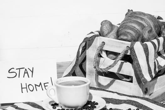 Coffee cup and buttered fresh French croissant on wooden crate. Food and breakfast concept. Morning message “stay home” on white board