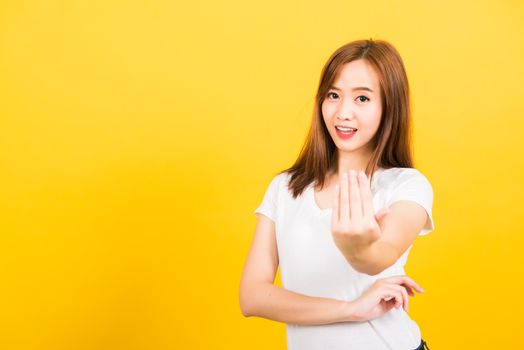 Asian happy portrait beautiful cute young woman teen smile standing wear white t-shirt making gesture hand inviting to come looking to camera isolated, studio shot on yellow background with copy space