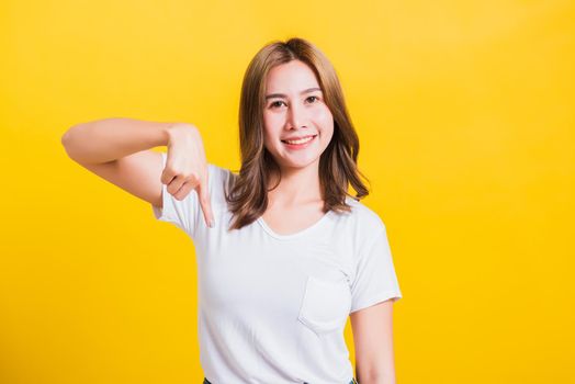 Asian Thai happy portrait beautiful cute young woman standing wear t-shirt makes gesture two fingers point below down and looking to camera, studio shot isolated on yellow background with copy space