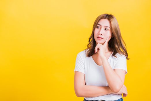 Portrait Asian Thai beautiful young woman wearing white t-shirt standing chin handle relaxed thinking about something about the question studio shot, isolated on yellow background with copy space