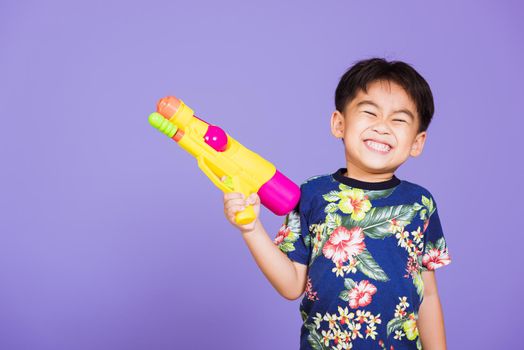 Happy Asian little boy holding plastic water gun, Thai kid funny hold toy water pistol and smiling, studio shot isolated on purple background, Thailand Songkran festival day national culture party