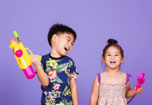 Two Happy Asian little boy and girl holding plastic water gun, Thai children funny hold toy water pistol and smile, studio shot isolated on purple background, Thailand Songkran festival day culture.