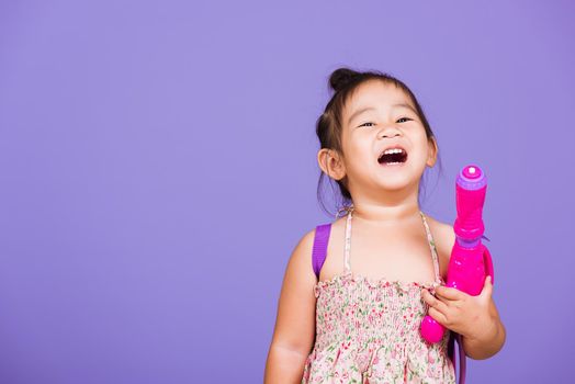 Happy Asian little girl holding plastic water gun, Thai child funny hold toy water pistol and smile, studio shot isolated on purple background, Thailand Songkran festival day national culture concept