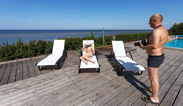 Photographer and nude model outdoors. Beauty, nudity and a healthy lifestyle. Beautiful nude woman lies on a sun lounger near the pool by the sea