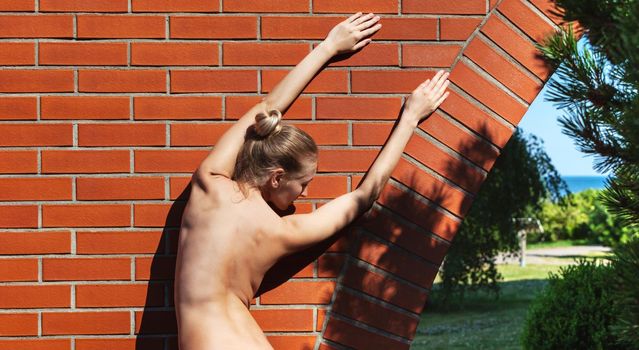 Young nude woman stands near a brick wall. Back view