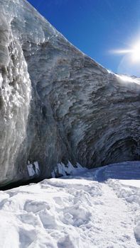 High ice wall in the mountains. The Bogdanovich Glacier. The black-and-white ice mixed. Grains of snow fly from the top of the glacier. The sun is shining. Lots of snow. Stones were frozen in places