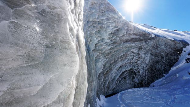 High ice wall in the mountains. The Bogdanovich Glacier. The black-and-white ice mixed. Grains of snow fly from the top of the glacier. The sun is shining. Lots of snow. Stones were frozen in places