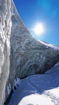 High ice wall in the mountains. The Bogdanovich Glacier. The black-and-white ice mixed. Grains of snow fly from the top of the glacier. The sun is shining. Lots of snow. Stones were frozen in places