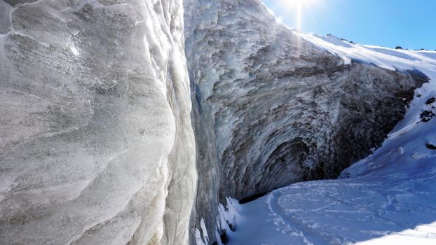 High ice wall in the mountains. The Bogdanovich Glacier. The black-and-white ice mixed. Grains of snow fly from the top of the glacier. The sun is shining. Lots of snow. Stones were frozen in places