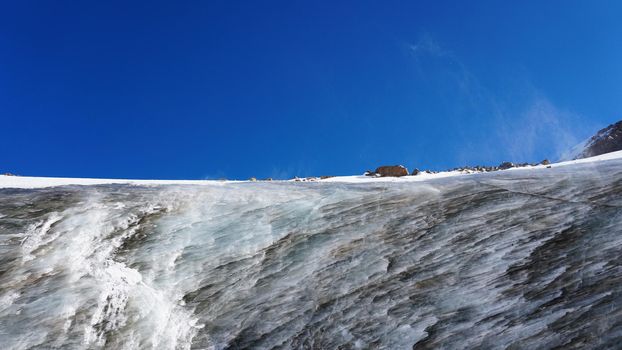 High ice wall in the mountains. The Bogdanovich Glacier. The black-and-white ice mixed. Grains of snow fly from the top of the glacier. The sun is shining. Lots of snow. Stones were frozen in places