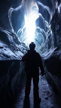 A climber with an ice pick stands at the exit of an ice cave. Inside Bogdanovich Glacier. High ice walls are sometimes covered with snow and stones lie. A flashlight is shining. Mountains of Almaty