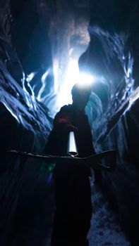A climber with an ice pick stands at the exit of an ice cave. Inside Bogdanovich Glacier. High ice walls are sometimes covered with snow and stones lie. A flashlight is shining. Mountains of Almaty