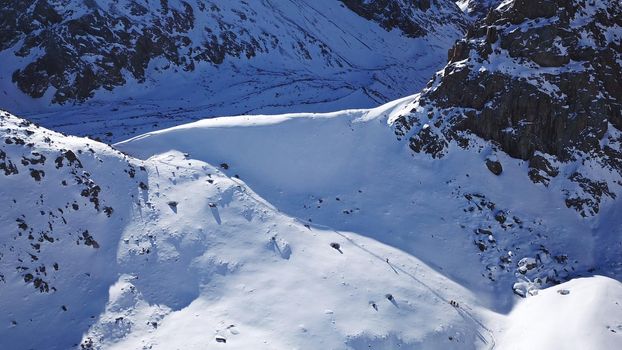 A group of climbers is walking in the mountains. Top view of the trail and the people. Snow-capped peaks, steep cliffs and precipices. A glacier is visible in the distance. Lots of snow. Almaty