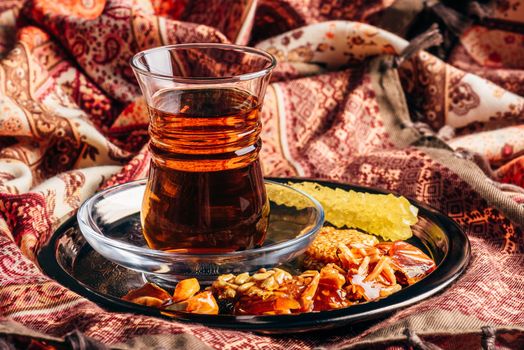 Black tea in armudu glass with candies with different nuts and crystal sugar on metal tray over tablecloth