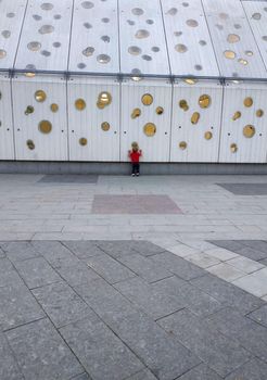 A small boy in a red T-shirt looks out the round window of the building. A building with many round windows. Light in the window.