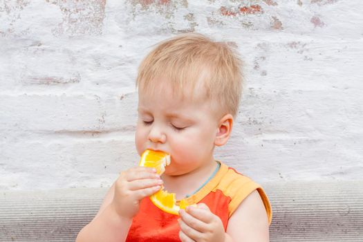 Portrait of child. Cute boy posing and eating a delicious orange. The emotions of a child. Portrait of child. Boy on the background of a brick wall.          