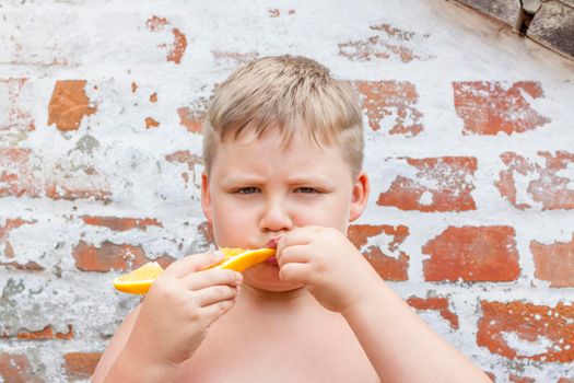 Portrait of child. Cute boy posing and eating a delicious orange. The emotions of a child. Portrait of child. Boy on the background of a brick wall.          