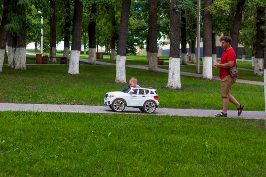 A small child is driven by a father in a children's car using a remote control. A man drives a car in which a child is sitting. 