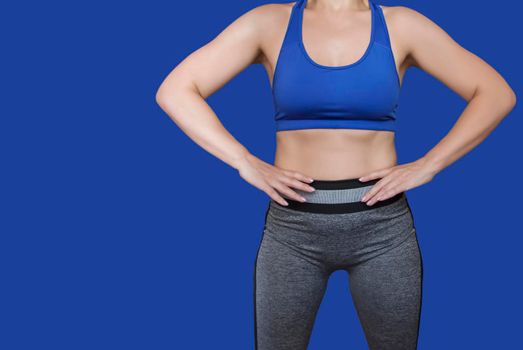 Fitness girl in a top and leggings points to her athletic figure. The Concept Of Weight Loss. Model on a blue background. A slender Young Lady Holds her hands at her waist. Free Space For Text.