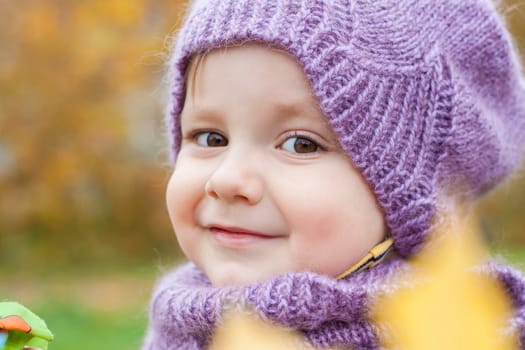 A child in a purple, knitted hat and scarf. Portrait of a child on the background of autumn trees.