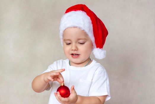 Portrait of a cute little boy in a white t-shirt and Santa Claus hat. A boy admires a red Christmas ball. Children's emotions. Christmas and new year. The concept of an advertising banner.