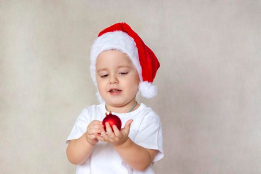 Portrait of a cute little boy in a white t-shirt and Santa Claus hat. A boy admires a red Christmas ball. Children's emotions. Christmas and new year. The concept of an advertising banner.  