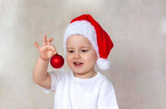 Portrait of a cute little boy in a white t-shirt and Santa Claus hat. A boy admires a red Christmas ball. Children's emotions. Christmas and new year. The concept of an advertising banner