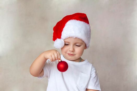 Portrait of a cute little boy in a white t-shirt and Santa Claus hat. A boy admires a red Christmas ball. Children's emotions. Christmas and new year. The concept of an advertising banner. 