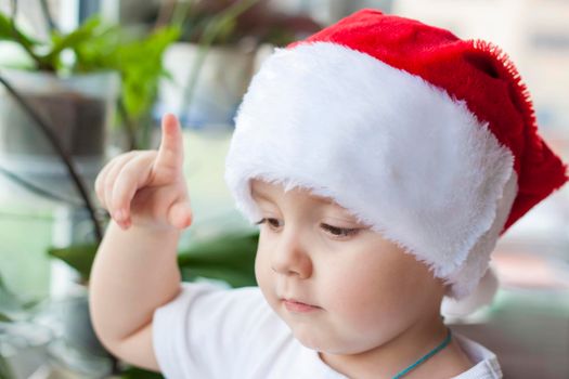 Portrait of a cute little boy in a white t-shirt and Santa Claus hat. Children's emotions. Christmas and new year. The child looks out the window. The concept of an advertising banner.