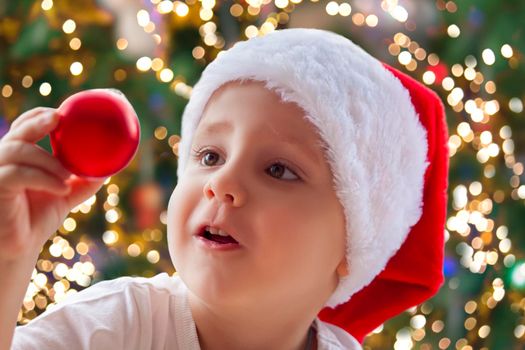 Child boy takes out of the box Christmas toys to dress up the tree. The Christmas tree is lit up and the baby looks at the Christmas toys with pleasure and joyful emotions. Christmas and new year. 