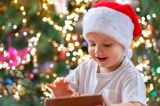 Child boy takes out of the box Christmas toys to dress up the tree. The Christmas tree is lit up and the baby looks at the Christmas toys with pleasure and joyful emotions. Christmas and new year.