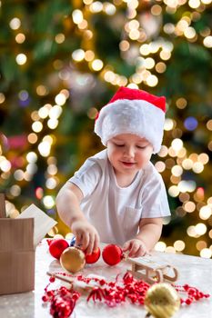 Child boy takes out of the box Christmas toys to dress up the tree. The Christmas tree is lit up and the baby looks at the Christmas toys with pleasure and joyful emotions. Christmas and new year. 