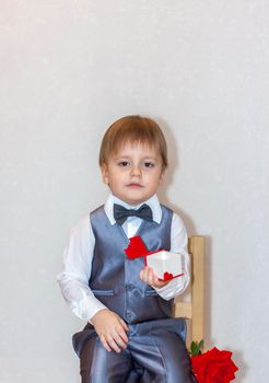 A little boy holds and hands over a red box, a Valentine's Day theme concept. Portrait of a cute boy in a suit with a bow tie. Valentine's Day.