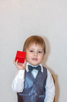 A little boy holds and hands over a red box, a Valentine's Day theme concept. Portrait of a cute boy in a suit with a bow tie. Valentine's Day.