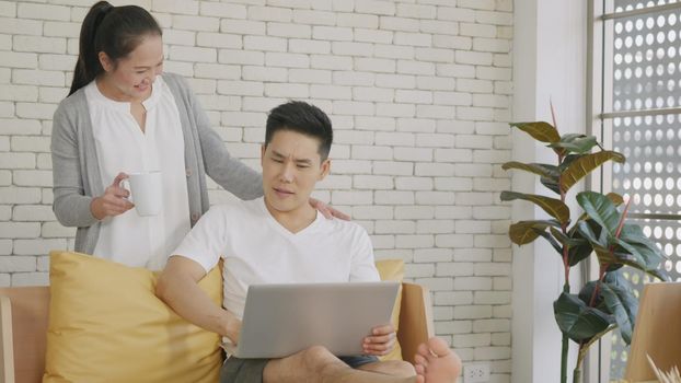 Happy Asian beautiful family couple husband and wife laughing sitting on sofa in the living room working with laptop computer at home. Woman brings coffee to the man during work