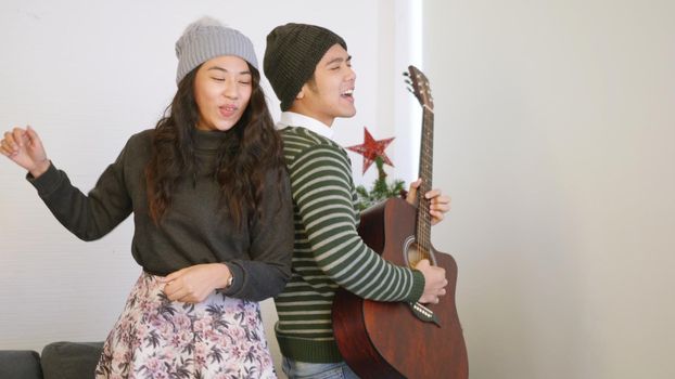 Happy Asian beautiful young family couple husband and wife on in home living room, man and woman playing the guitar, singing song and dancing together enjoying great time in living room, 