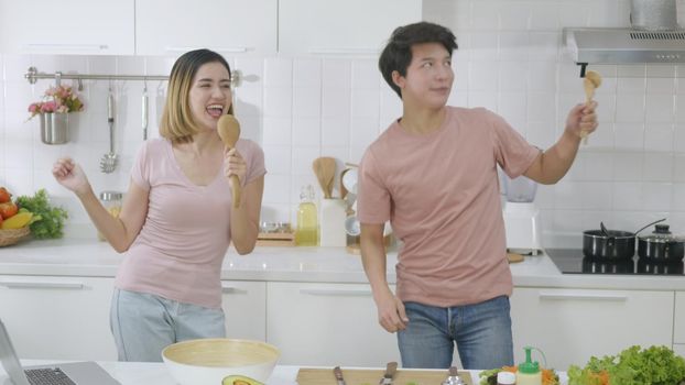 Happy Asian beautiful family couple husband and wife singing in kitchenware microphones in kitchen together having fun dance listen music at home. Two people dancing