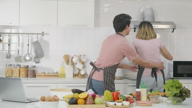 Happy Asian beautiful family couple husband and wife cooking vegetable salad in kitchen together at home. The man sneaked in to pretend to tickle the waist woman. healthy food concept